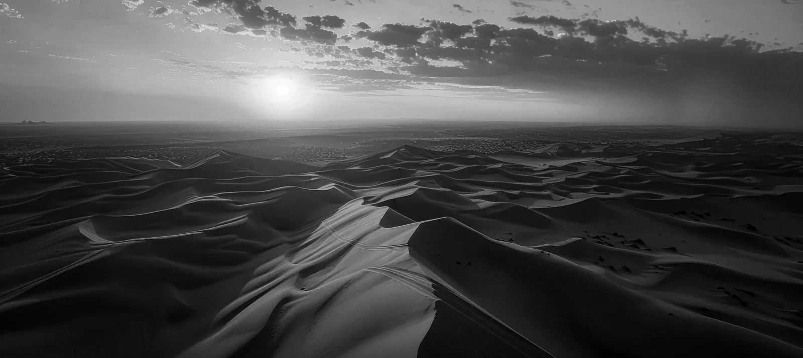 Aerial black-and-white panorama of vast sand dunes beneath a cloud-streaked sky